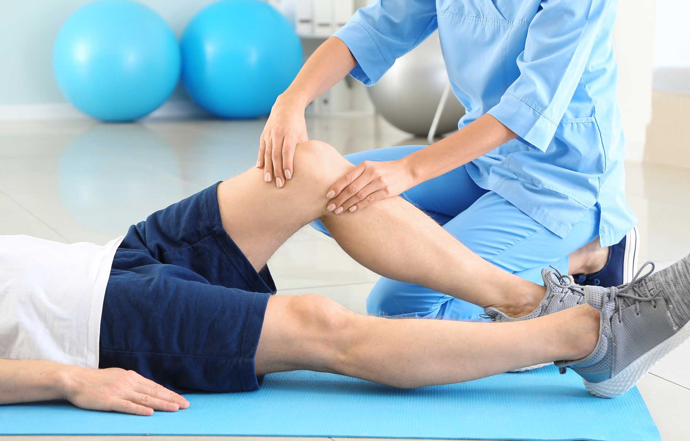 Un fisioterapeuta con bata azul ayuda a una persona tumbada en una colchoneta sujetando y apoyando suavemente su rodilla doblada durante una sesión de rehabilitación o terapia. La fisioterapia general está orientada a tratar el dolor, recuperar la movilidad y mejorar tu calidad de vida. Fisioterapia general