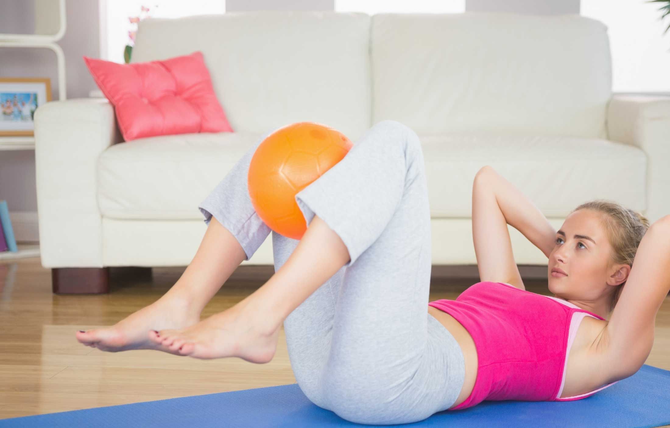 Una mujer con ropa de gimnasia hace ejercicios abdominales sobre una esterilla de yoga, sujetando una pelota naranja entre las rodillas, con un sofá blanco y una almohada rosa de fondo. Fisioterapia suelo pélvico