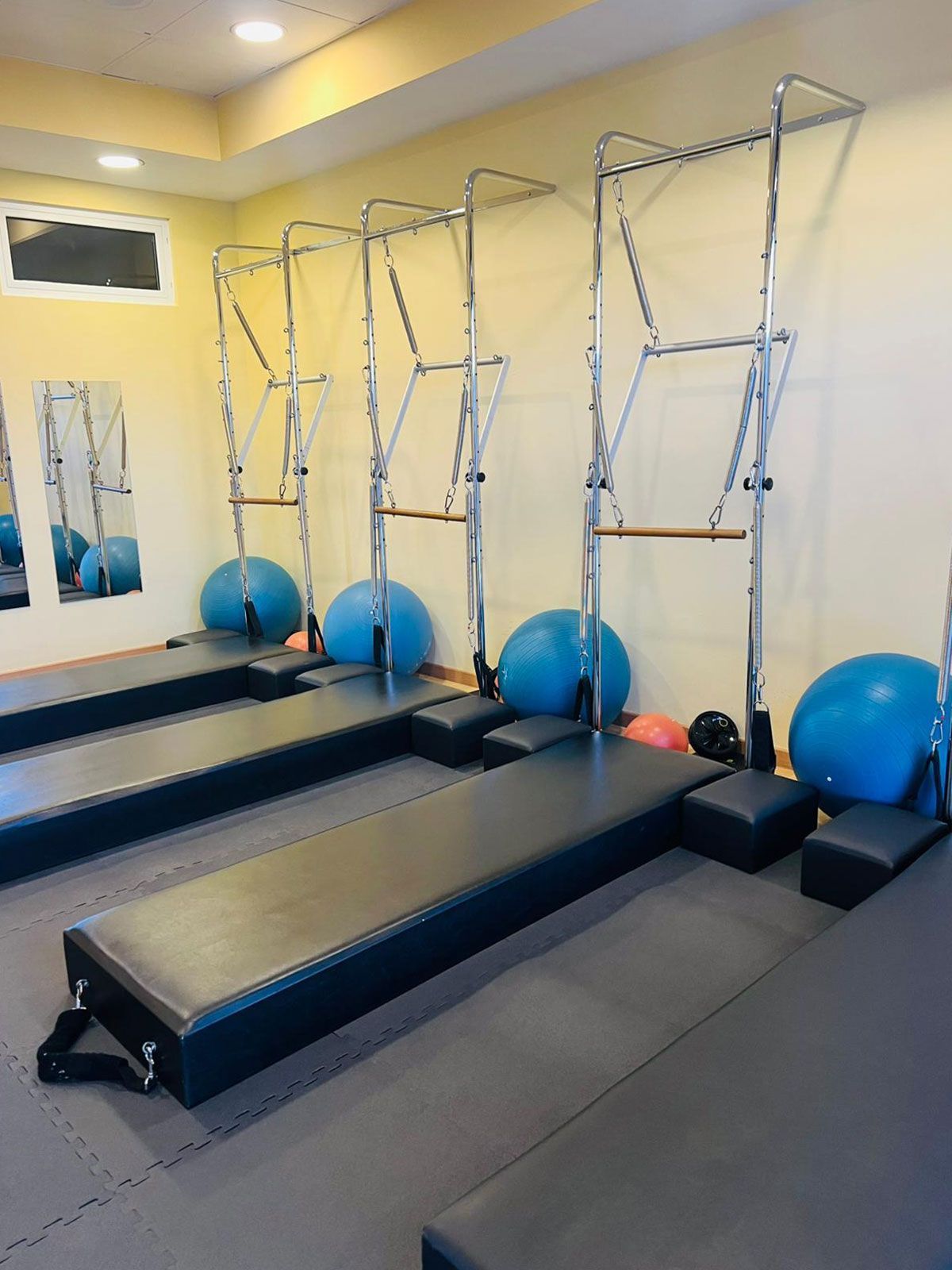 Un estudio de Pilates con tres torres de Pilates, bancos acolchados negros, pelotas de ejercicio azules y rosas, pequeños bloques negros y un espejo de pared que refleja el equipo. La sala tiene paredes amarillas y una iluminación tenue.