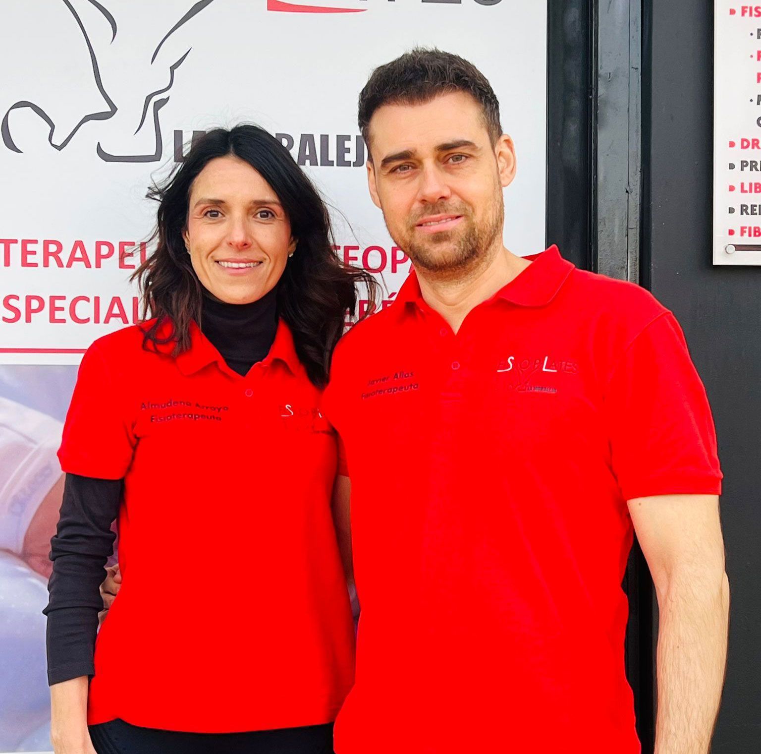 Dos personas con polos rojos a juego, de pie, una al lado de la otra, sonríen a la cámara delante de un cartel de una clínica con texto y gráficos médicos de fondo.